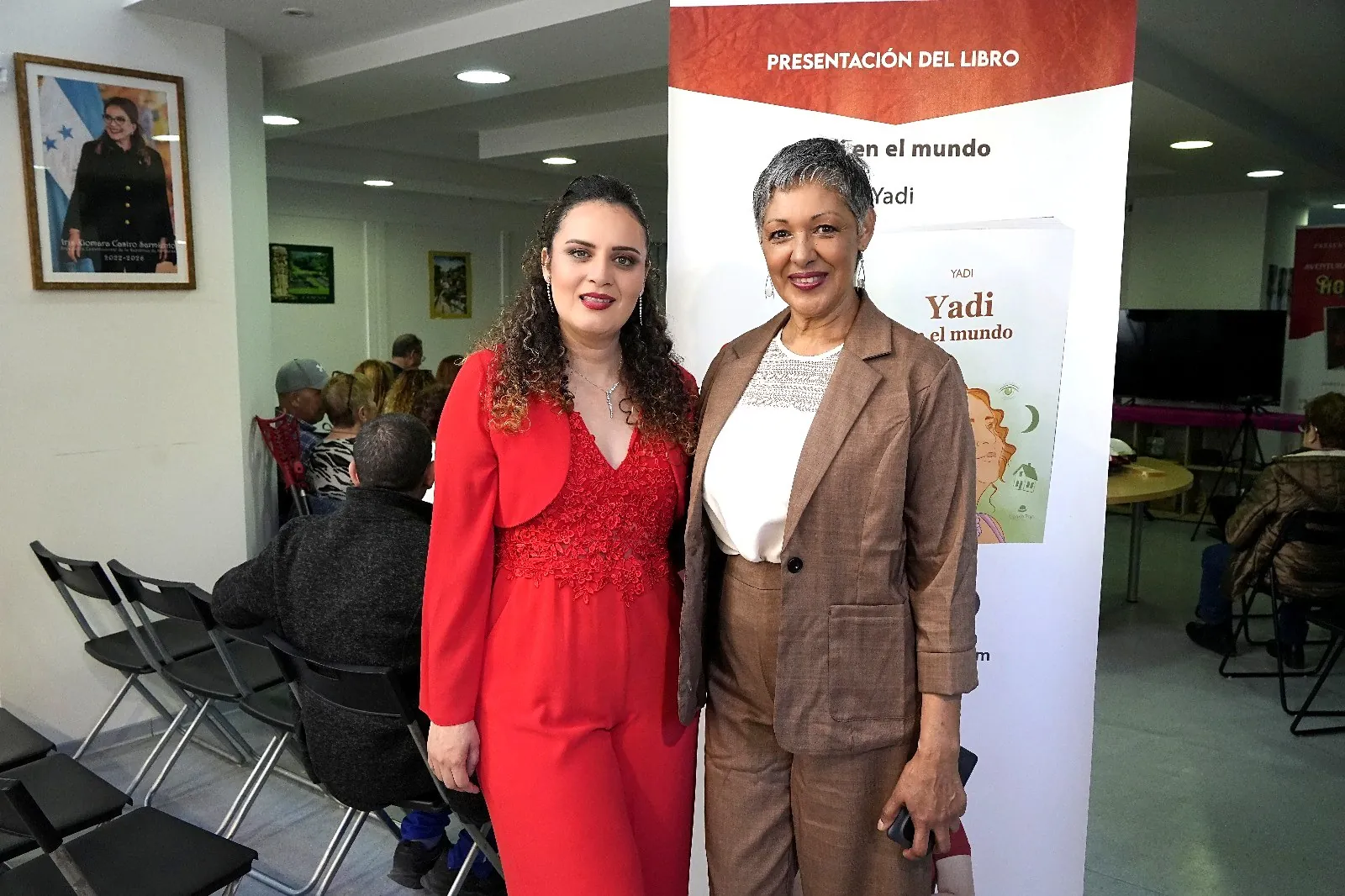 Yadi Villanueva junto a una invitada ante el cartel de la presentación del libro en el Consulado de Honduras